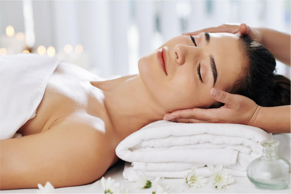woman getting her face massaged whilt having eyes closed and head resting on folded towels in a white setting