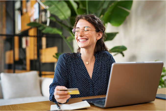 Smiling woman with glasses making an online booking on her laptop whilst holding a credit
			card